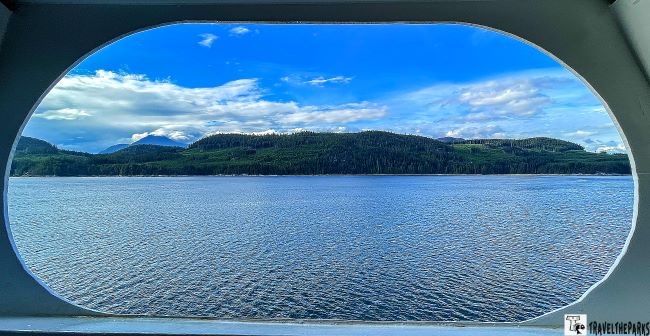 View of the inside passage and tree-lined shore through a ship porthole window frame on our ultimate Alaska cruise of the inside passage