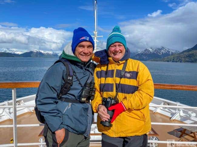 Two people in winter clothing on a boat deck with snow-capped mountains in the background on a tour of Glacier Bay National Park