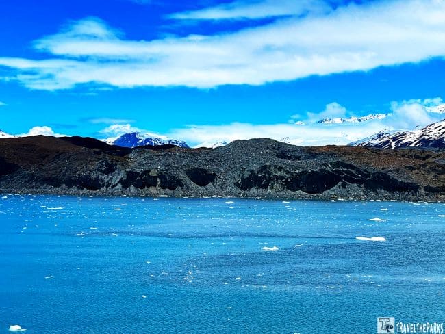 Grand Pacific Glacier in Tarr Inlet with icebergs and rocky shoreline under blue sky and snowy mountains.