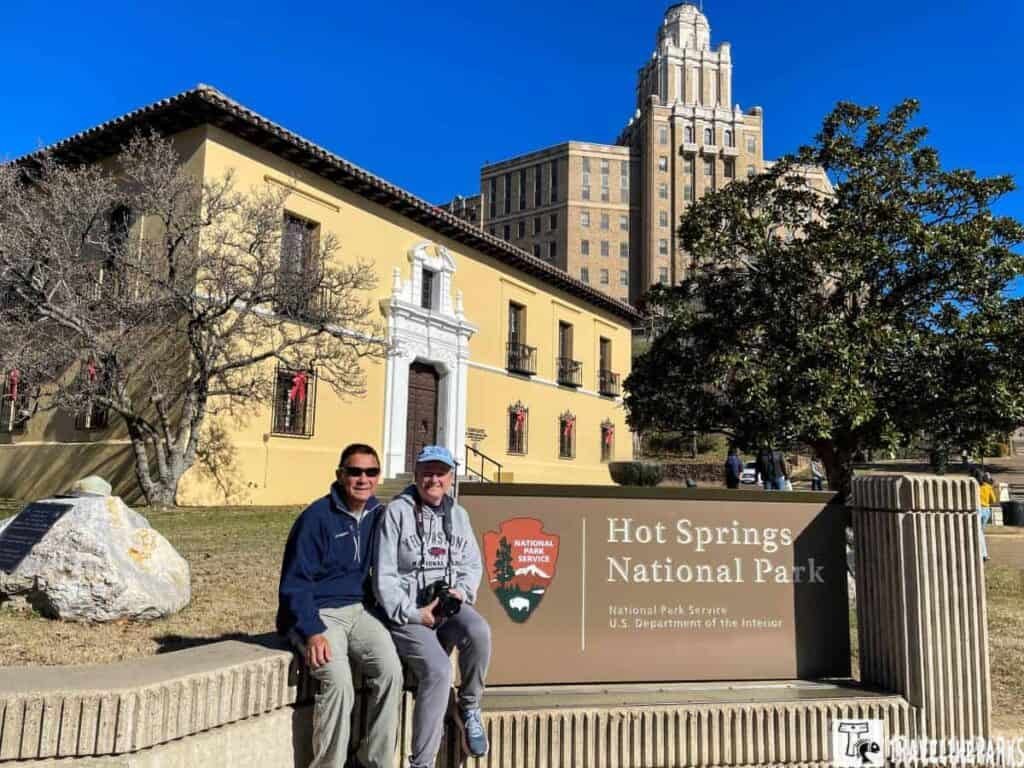 Two people sitting in front of Hot Springs National Park with historic buildings and trees.