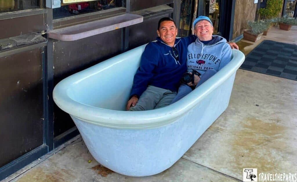 Two people sitting in a white standalone bathtub on a concrete floor outside a building.