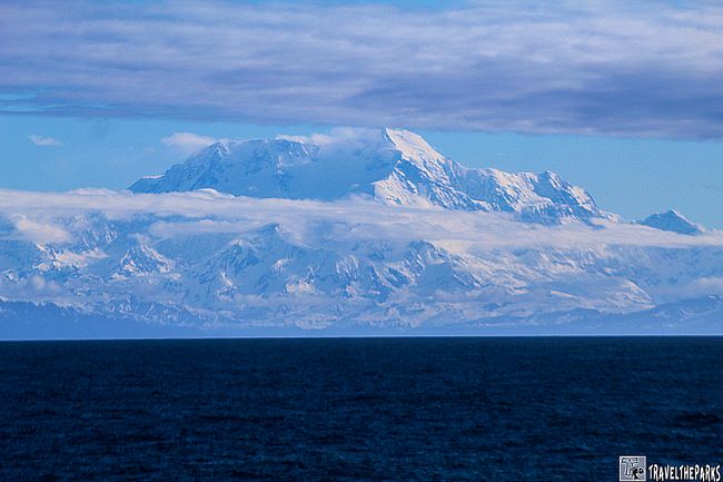 A snow-covered Mount St. Elias with clouds hovering above, seen across a deep blue ocean.