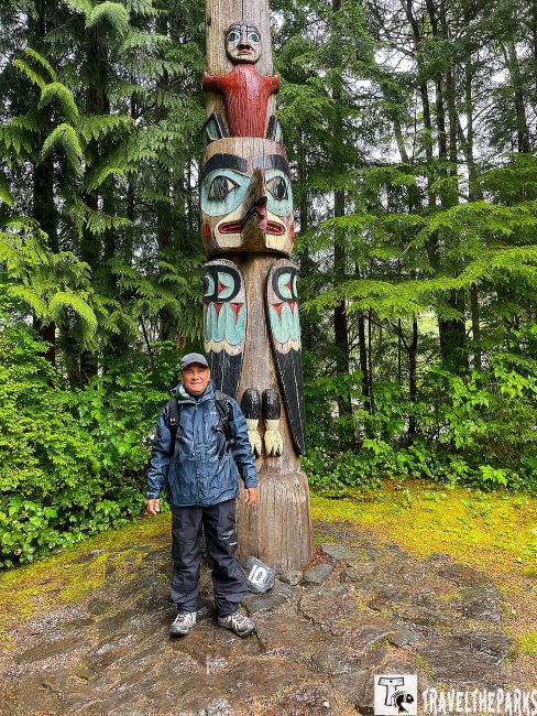 Totem pole at Totem Bight State Park on an inside passage cruise surrounded by dense greenery with a person standing in front wearing a rain jacket.