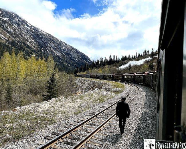 White's Pass Summit Train t on curved tracks with mountains and trees in the background; conductor walks beside the tracks.