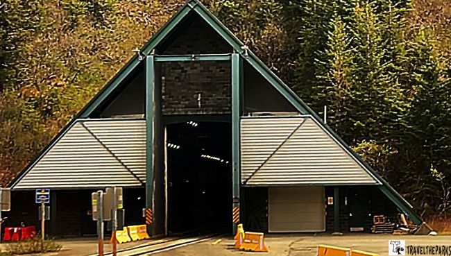Triangular Anton Anderson Memorial Tunnel entrance with teal structure and orange barriers in a forested area.