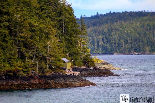 A coastal landscape with a rustic cabin by a forested shoreline and expansive water.