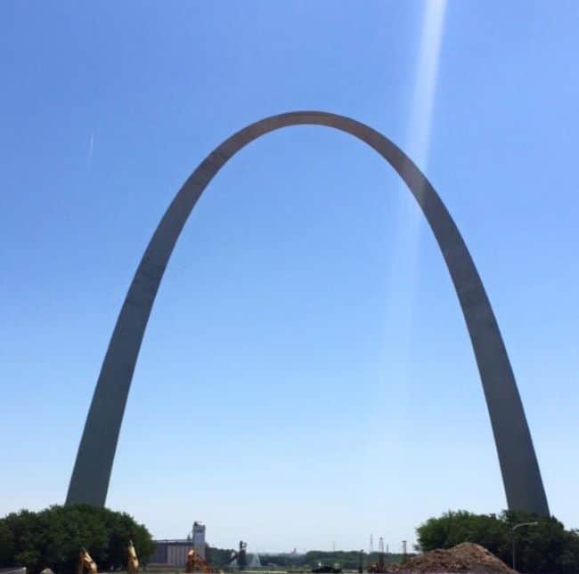 The image shows the Gateway Arch, a curved stainless steel monument situated in a clear blue sky. The arch rises gracefully from a green landscape at its base with a few scattered buildings and trees visible in the background—a must-see landmark on any Midwest road trip.