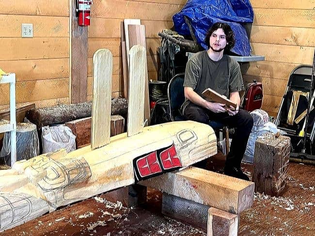 Male Totem carver sitting near wooden sculpture at Saxman Village Totem Park