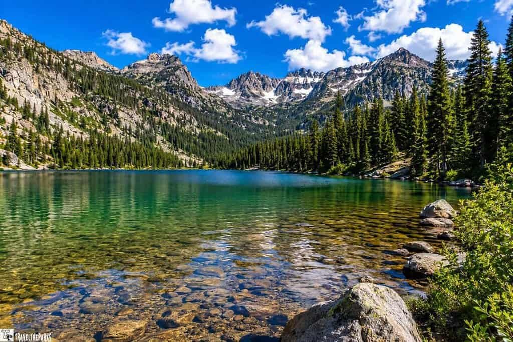 A serene mountain lake with clear water reflecting the surrounding dense forest. Rocky shorelines frame the sides of the image, and a fallen log rests at the water's edge on the right. The sky is clear and blue, allowing abundant sunlight to illuminate the scene.