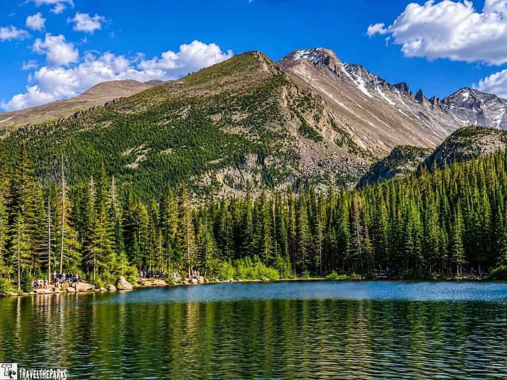 A serene mountain landscape with a clear lake in the foreground, dense evergreen trees lining the shore, and rugged, snow-capped peaks against a partly cloudy sky in the background. The trees' reflection is visible in the calm water of Bear Lake in Rocky Mountain National Park.