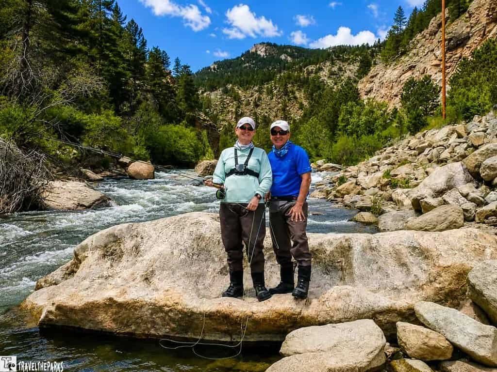 Big Thompson River Guided Fishing: Two people in fishing attire stand on rocks beside a flowing river. They wear hats and sunglasses, holding fishing rods, surrounded by lush greenery.