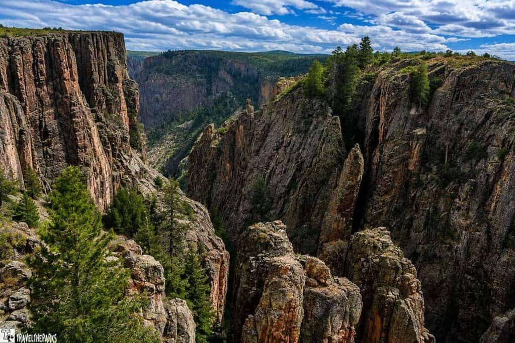 A dramatic landscape featuring a rugged canyon with steep, rocky cliffs. The scene is lush with greenery, as trees and shrubs cling to the rocky surfaces. Overhead, a cloudy sky creates a stark contrast with the vibrant natural elements below—perfect for a picturesque Colorado road trip to the Black Canyon of the Gunnison.
