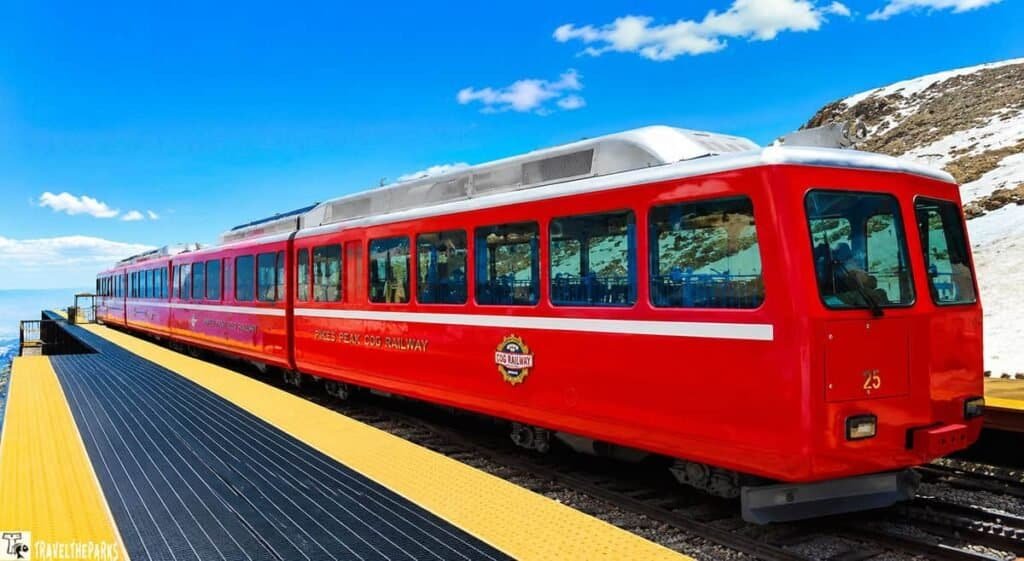 A red cog railway train on a yellow platform at Pikes Peak, set against a blue sky with scattered clouds and snow-capped mountains in the background.

