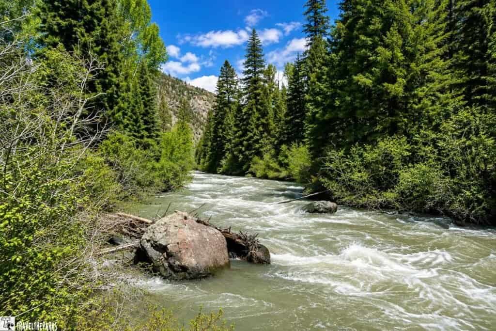 A river flows through a forested area with greenery along the banks, large boulders, and a clear blue sky with clouds.

