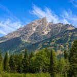 A scenic view of jagged mountains surrounded by dense green forests under a bright blue sky with wispy clouds.
