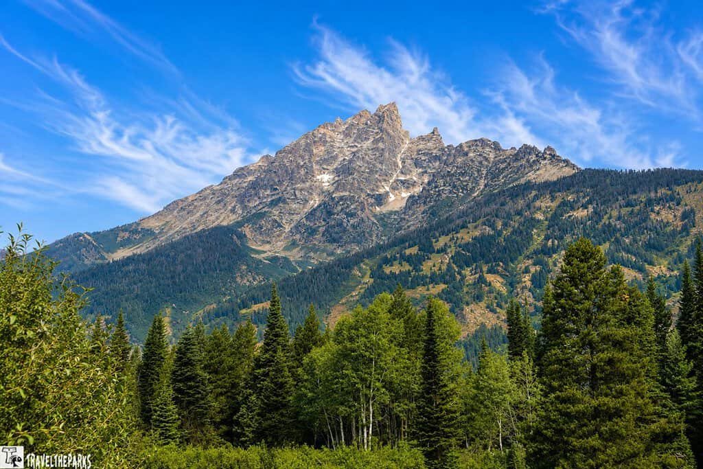 A scenic view of jagged mountains surrounded by dense green forests under a bright blue sky with wispy clouds.
