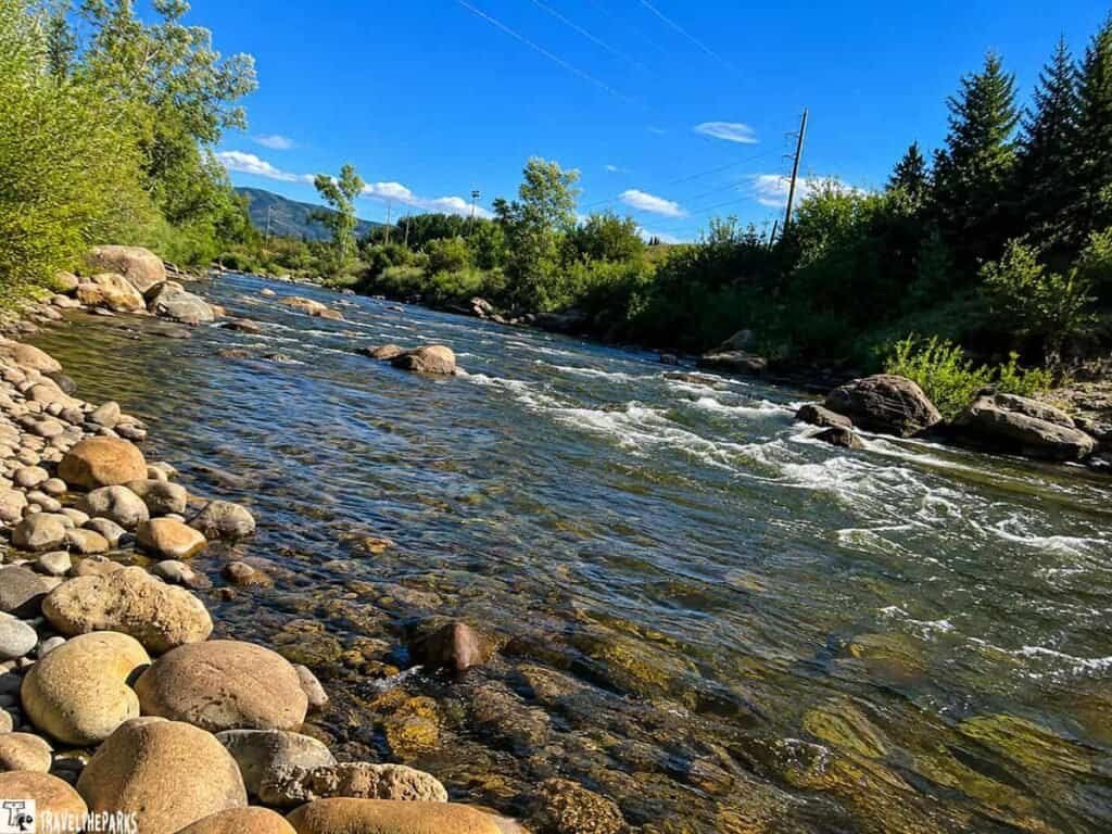 A peaceful river flows through a rocky bed, surrounded by greenery and trees under a clear blue sky.

