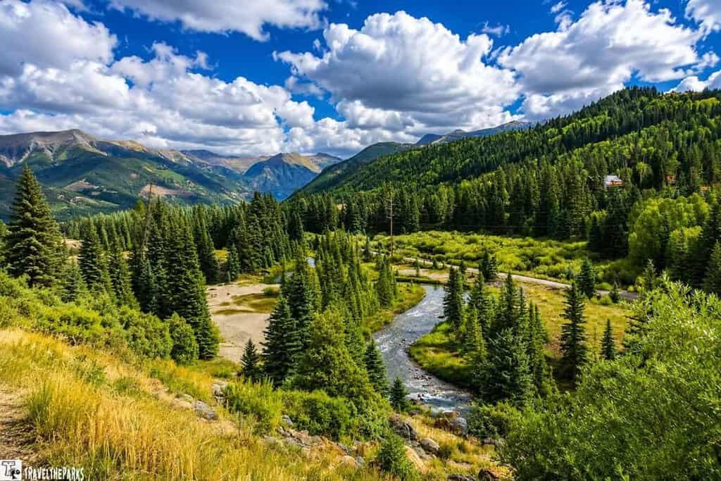A scenic view of a lush, green forested valley with a winding river flowing through it. Majestic mountains can be seen in the background under a sky filled with clouds. This picturesque landscape is perfect for any 10 Day National Park Colorado Trip Itinerary, showcasing tall pine trees and serene natural beauty.