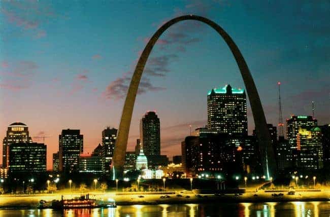 The Gateway Arch is in the center, the old St. Louis Courthouse below it and many tall skyscrapers behind. There is a riverboat on the bank.