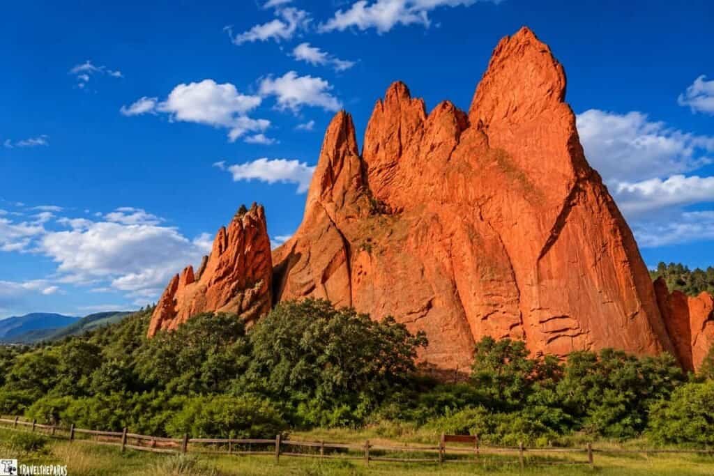 A towering red rock formation stands against a bright blue sky, with sunlight streaming down from above. Green vegetation grows at the base, contrasting with the rugged, reddish-brown surface of the rocks.
