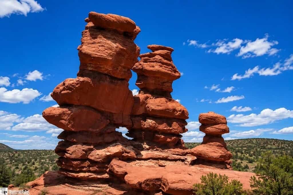 A set of reddish-brown rock formations with a hole at the base, surrounded by green hills and under a bright blue sky with clouds.