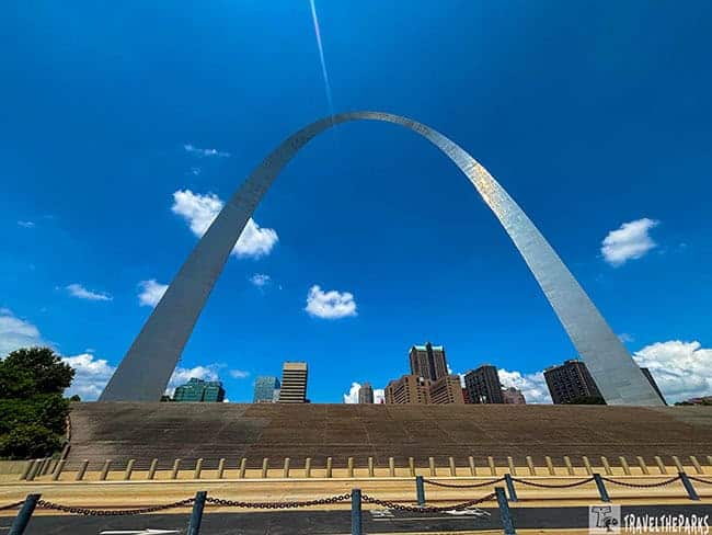 A towering metallic arch against a bright blue sky with clouds, overlooking the St. Louis skyline.