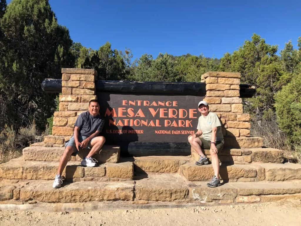 Two people sitting by the Mesa Verde National Park entrance sign.