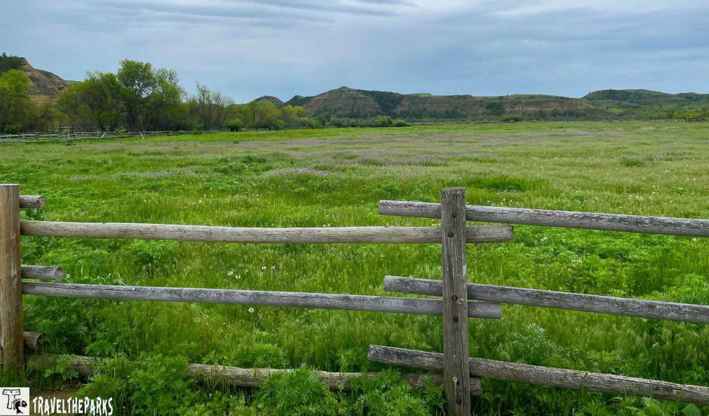 A serene countryside landscape featuring a wooden fence in the foreground, separating a lush, green field. Distant rolling hills and a partly cloudy sky add depth to the scene. Perfect for a Midwest national parks road trip