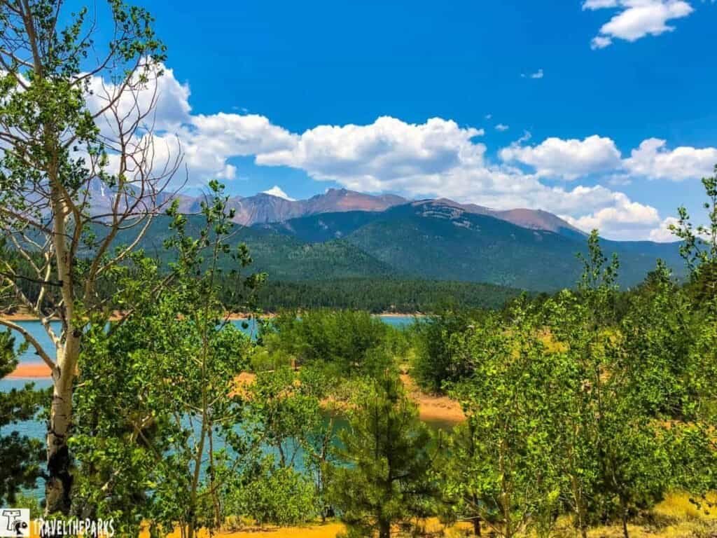 A scenic view of Crystal Lake Reservoir surrounded by green trees and distant mountains under a blue sky with white clouds.

