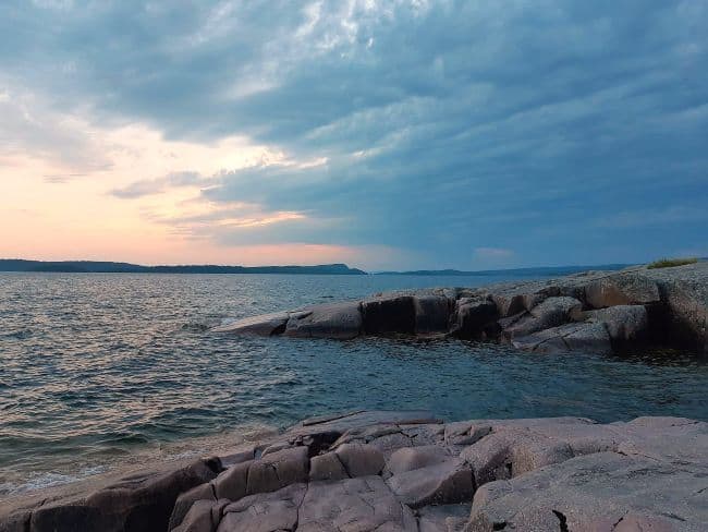 A rocky shoreline on Isle Royale at sunset, with calm waters and cloudy skies featuring pink and orange hues.