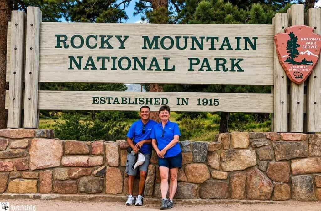 wo people wearing blue shirts pose in front of the Rocky Mountain National Park sign, which reads "Established in 1915." They are standing close together and smiling, with the wooded park area visible in the background.