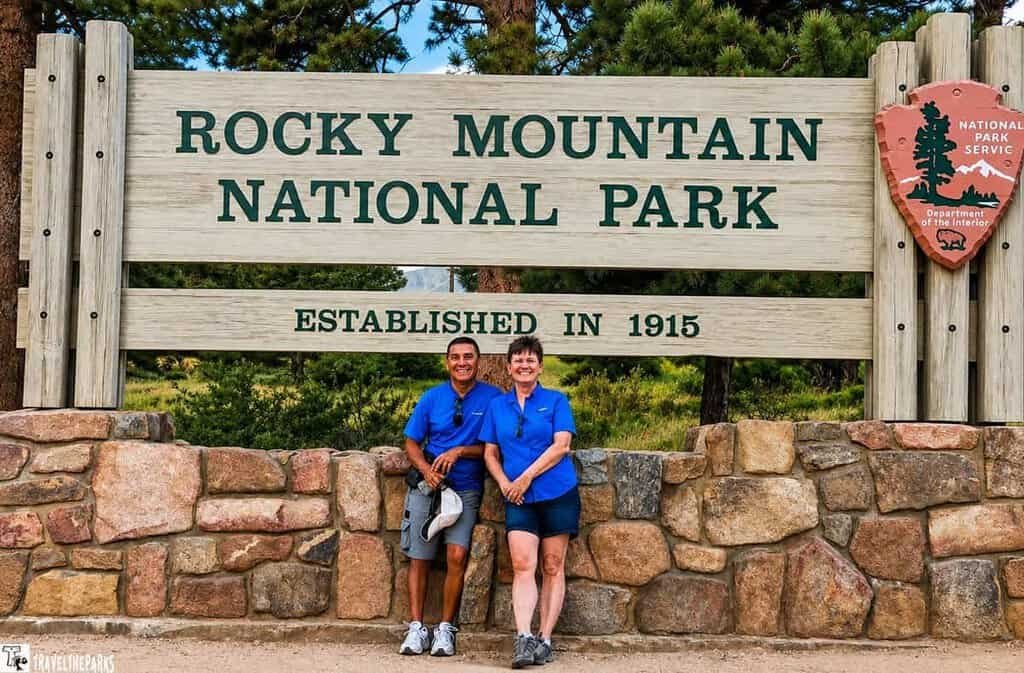 wo people wearing blue shirts pose in front of the Rocky Mountain National Park sign, which reads "Established in 1915." They are standing close together and smiling, with the wooded park area visible in the background.