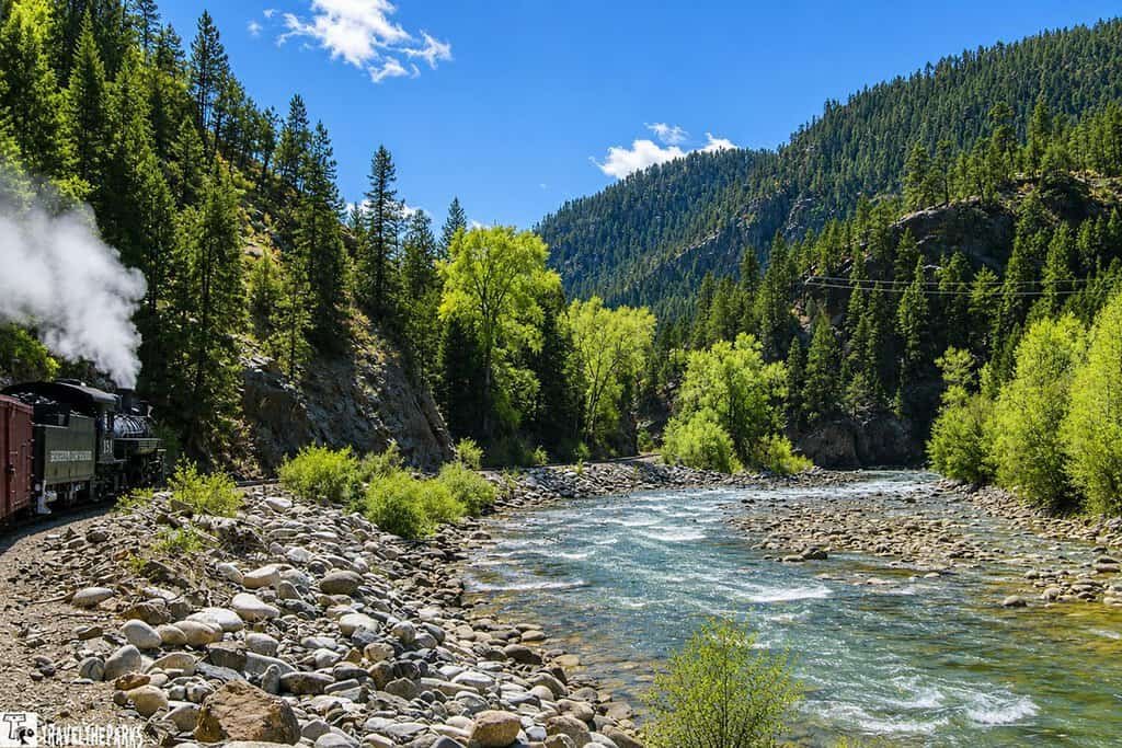 A steam locomotive runs alongside a rocky riverbank, surrounded by lush greenery and steep mountains under a blue sky.

