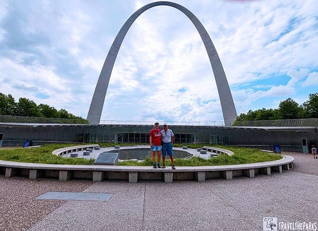 Two people pose in front of the Gateway Arch on a circular stone platform, surrounded by grass under a partly cloudy sky.