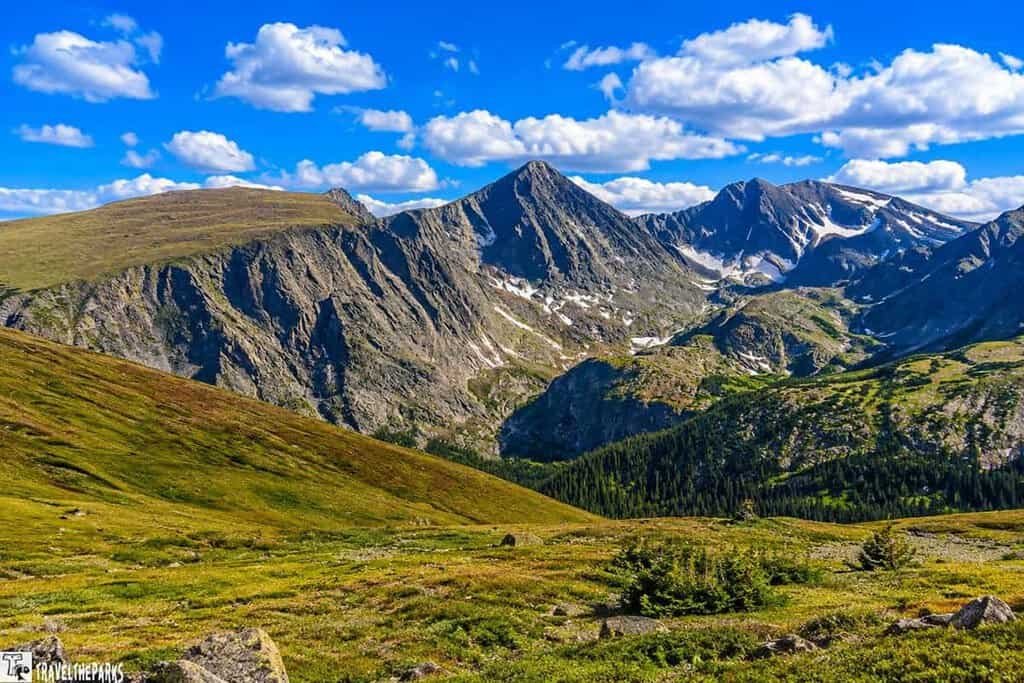 Mountain landscape with grassy foreground and rocky peaks under a blue sky with clouds.