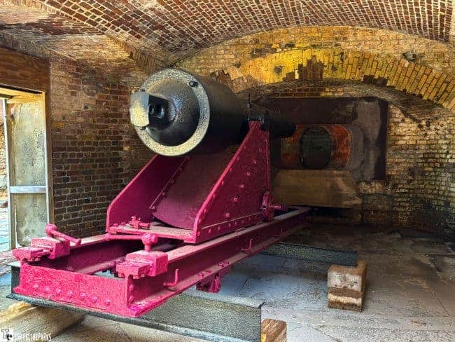 A large, old-fashioned black cannon mounted on a sturdy red base is displayed indoors against a backdrop of aged brick walls. The cannon appears well-preserved and is situated on a stone floor, showcasing historical military technology.
