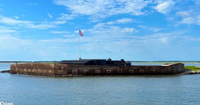 A historic fort made of brick and stone, Fort Sumter National Monument, sits by the water's edge. Several flags, including the U.S. flag, are flying from poles on top of the fort. The sky is clear with a few clouds, and the water appears calm near the rocky shoreline—an ideal stop on an East Coast road trip.