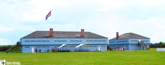 Two light-blue historic blockhouse buildings with a British flag, people in period dress, and outdoor tents on a grassy field.