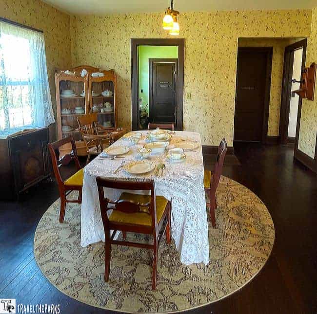 Vintage dining room with a lace-covered table set for a meal, surrounded by wooden chairs and a patterned rug.