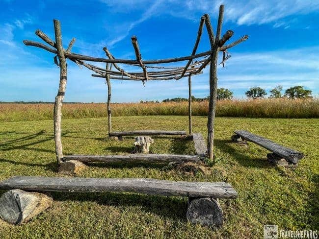 Rustic wooden frame structure with benches on a grassy field under a blue sky.
