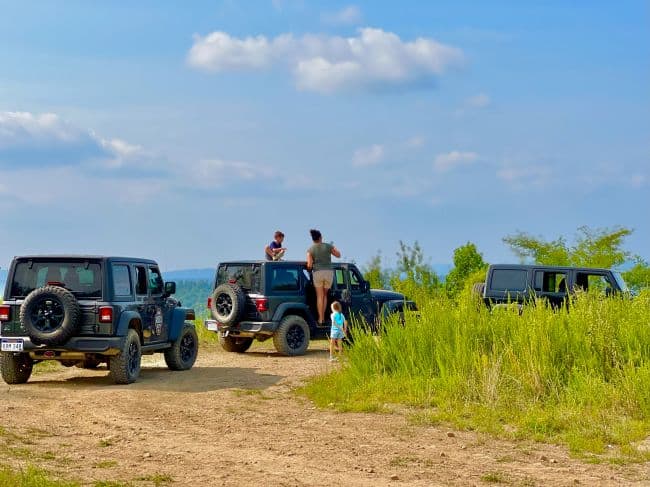 Three black Jeeps parked on a dirt path with people interacting near them, set against a scenic landscape with clouds and distant mountains.