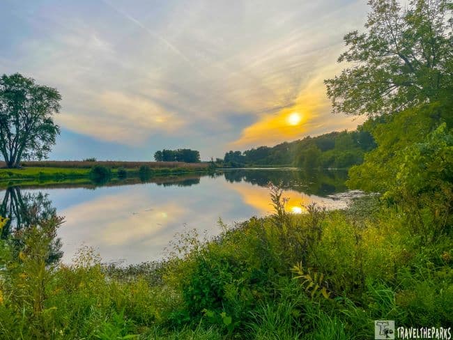 Prophetstown State Park landscape with calm water, lush greenery, and the sun setting or rising, casting a golden light.at Prophetstown State Park