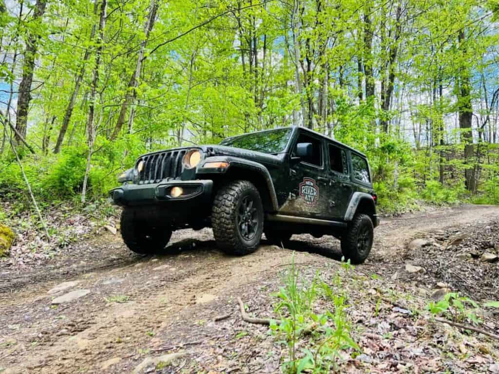 a black Jeep driving along a dirt trail in a forested area. The Jeep is angled slightly upward as it navigates the uneven, rocky terrain. The foreground features a mix of dirt and small rocks, with patches of green grass and leaves scattered around. Surrounding the Jeep, tall trees with bright green leaves suggest a spring or early summer setting. The sky above is partially visible with patches of blue. The Jeep has an emblem on the door that reads "New River ATV".