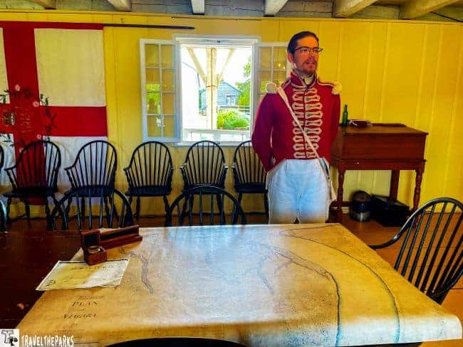 Historical reenactor in period uniform at Fort George dining hall stands by a table with a historical map, surrounded by antique chairs and a large flag.