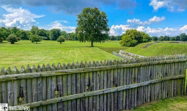 Wooden palisade fence with gaps, overlooking a grassy field and a river.