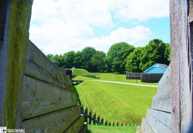 Wooden palisade walls of a historical fort overlook a grassy field and historical ravelin structures.