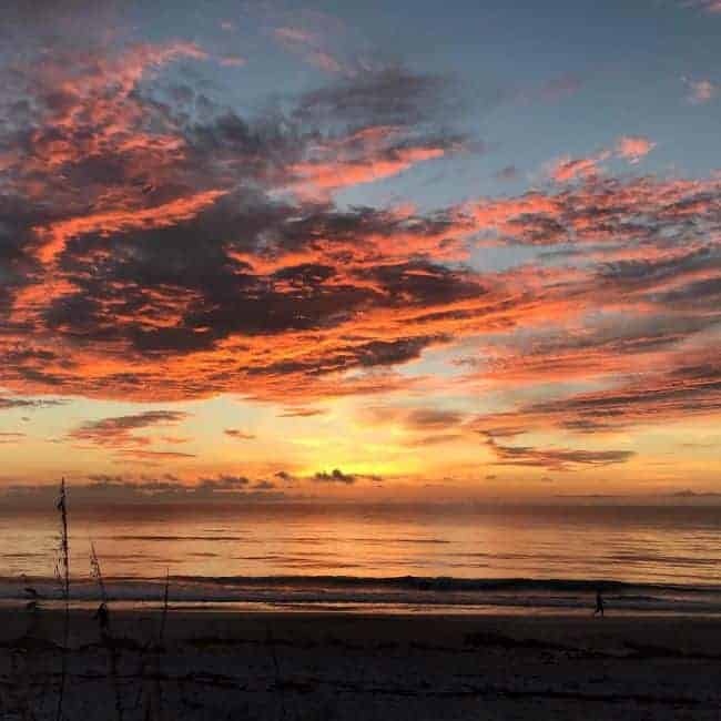 Vibrant sunrise over a calm river during one of the Sunshine State Orlando Day Trips, with colorful clouds reflecting on the water and a silhouetted person enjoying the peaceful morning on the shore.