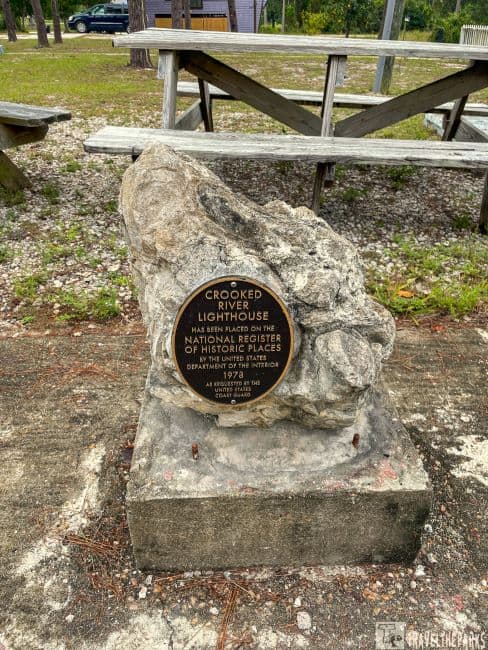 a weathered stone monument set on a square concrete base. Mounted on the stone is a circular plaque made of metal with engraved text. Surrounding the monument, the ground is covered with a mix of grass, small rocks, and dirt. Behind the monument, there are wooden picnic tables on a gravel surface. In the background, some trees and a parked vehicle are visible.