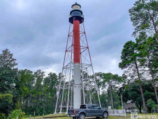Crooked River Lighthouse with a red and white conical frame and a black lantern, surrounded by trees and an overcast sky. A truck is parked at its base.