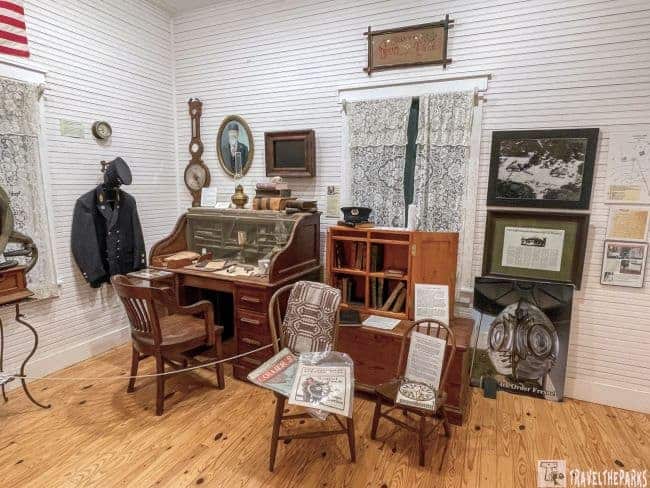 Museum room with nautical artifacts, including a navy jacket, roll-top desk, and Fresnel lens replica.